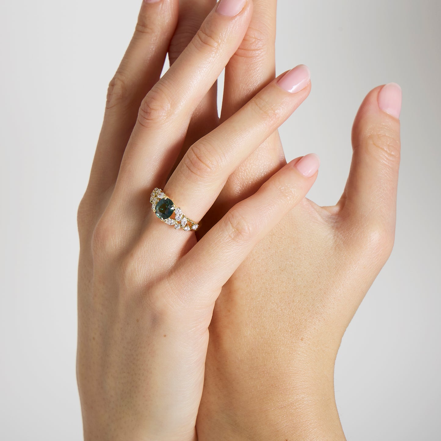 Gros plan sur deux mains claires ; l'une est ornée d'une bague Bonnot Paris Bague Saphir Teal • 2,07 ct ornée d'un saphir turquoise encadré de diamants taille marquise. Les ongles sont manucurés avec un vernis rose clair sur un fond sobre.