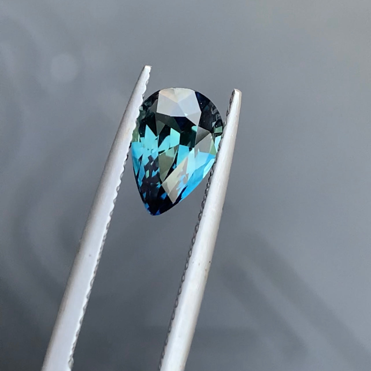 Une pierre précieuse multicolore en forme de poire pesant 2,06 carats aux teintes bleues et vertes est saisie par une pince métallique sur un fond neutre, mettant en valeur sa beauté enchanteresse rappelant le Saphir Teal de Bonnot Paris.