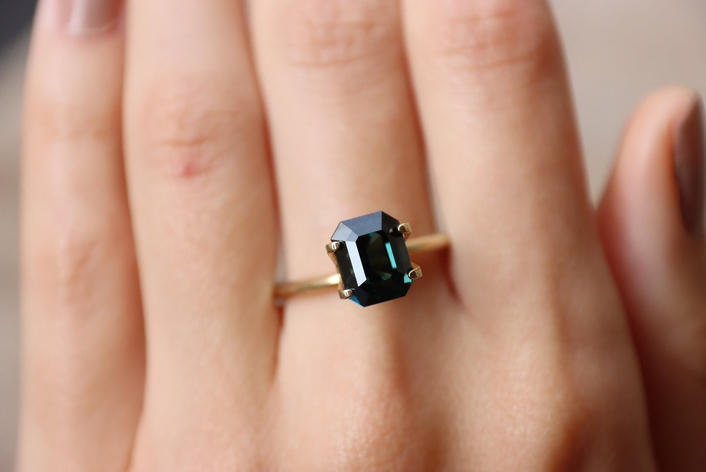 Une main porte la bague Saphir Teal en or 4,04 carats de Bonnot Paris, ornée d'une grosse pierre rectangulaire vert foncé. Les ongles sont manucurés naturellement.