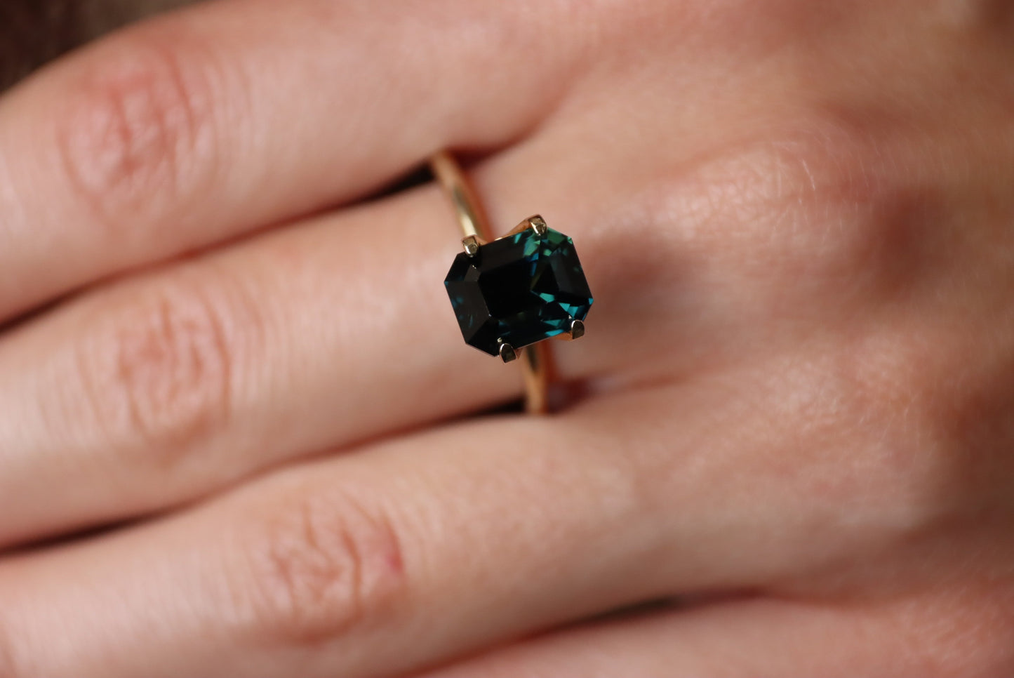 Une main porte le Saphir Teal · 4,04 carats de Bonnot Paris, orné d'une grande pierre rectangulaire vert foncé sertie sur or. Les doigts sont légèrement écartés, posés sur une surface.