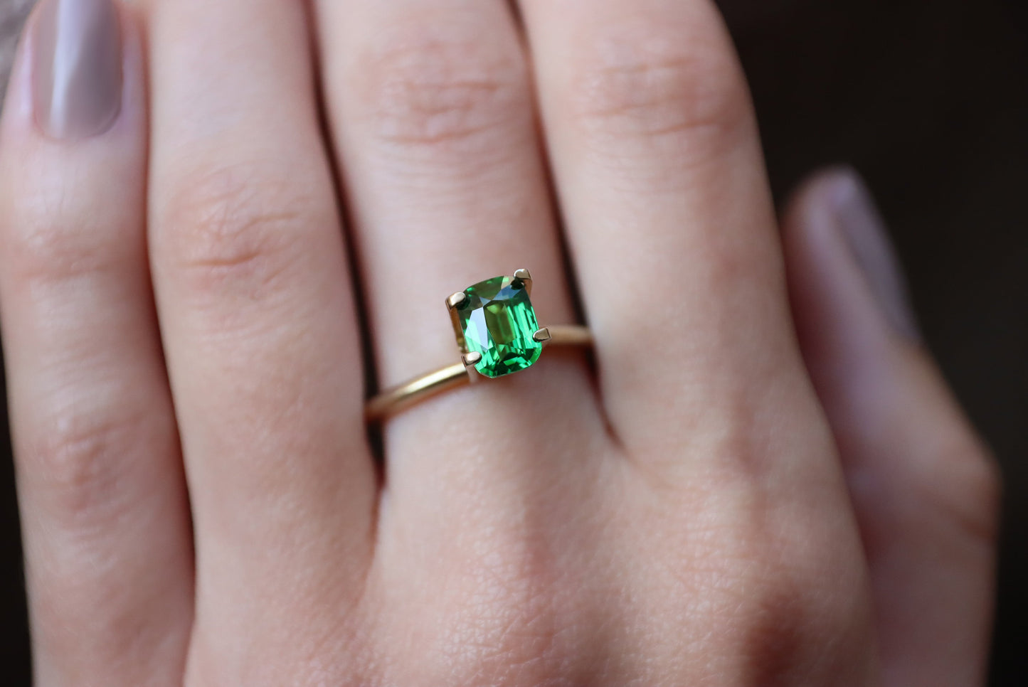 Gros plan d'une main portant la bague Bonnot Paris Grenat Tsavorite en or 1,54 carat ornée d'une pierre verte rectangulaire. Les doigts légèrement polis sont partiellement visibles sur un fond sombre et flou.