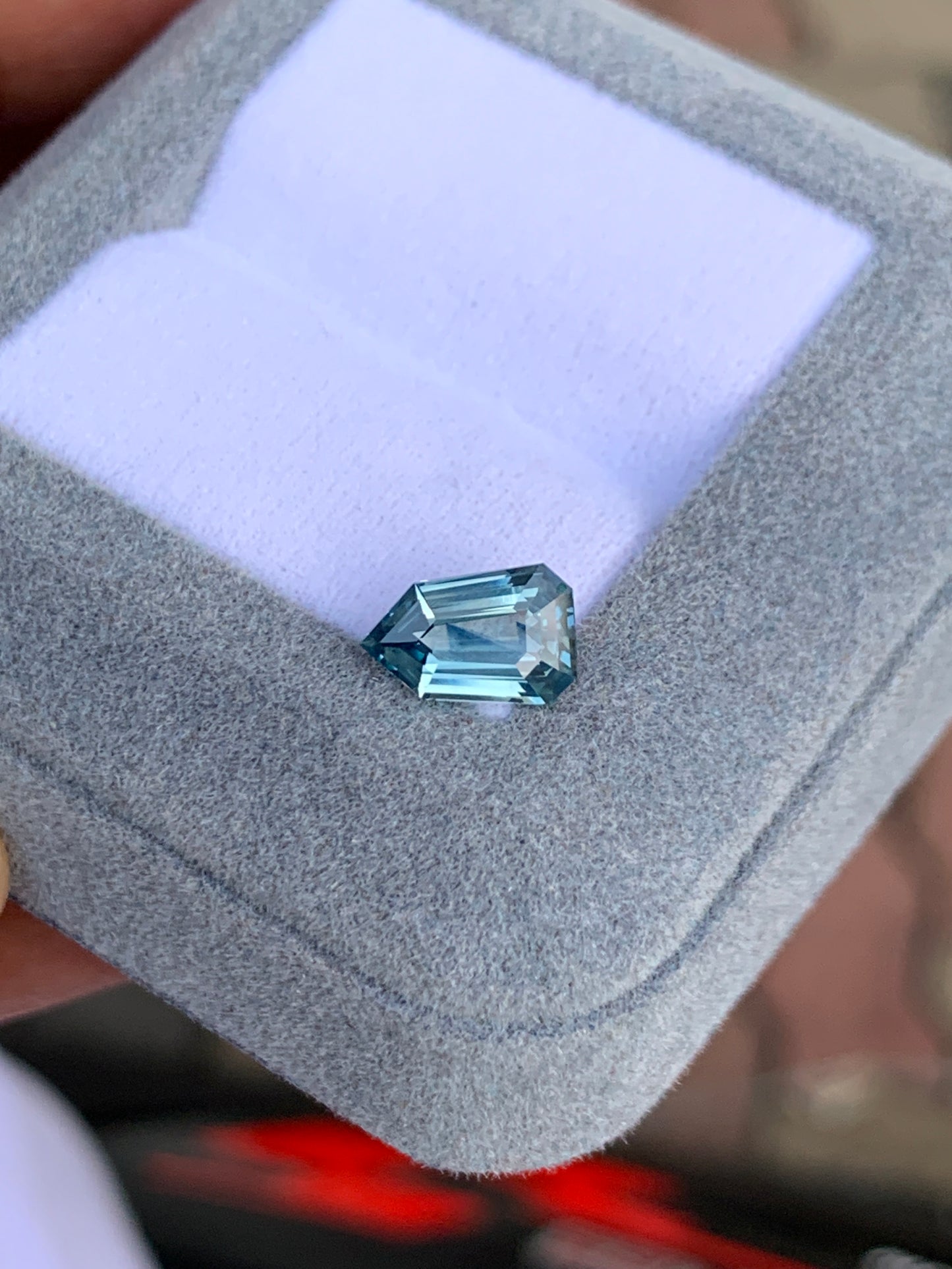 Le Saphir Teal de 1,65 carat de Bonnot Paris est présenté dans un écrin gris clair à l'intérieur blanc. Sa forme asymétrique aux multiples surfaces réfléchissantes met en valeur sa clarté et sa couleur sur un fond flou.