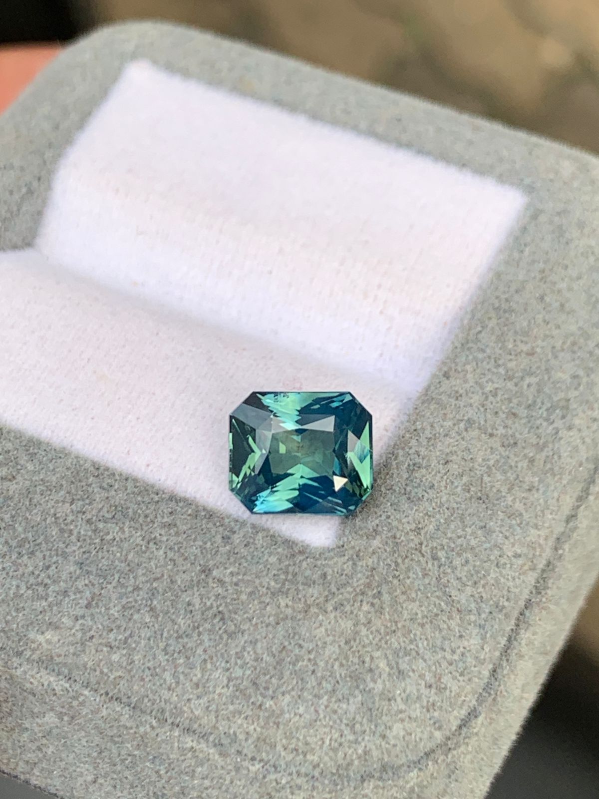 Le Saphir Teal • 3,10 carats de Bonnot Paris, une pierre précieuse rectangulaire facettée aux reflets bleus et verts, repose sur un coussin blanc dans un écrin gris ouvert. Pesant 3,10 carats, il reflète magnifiquement la lumière, soulignant sa clarté et son éclat.