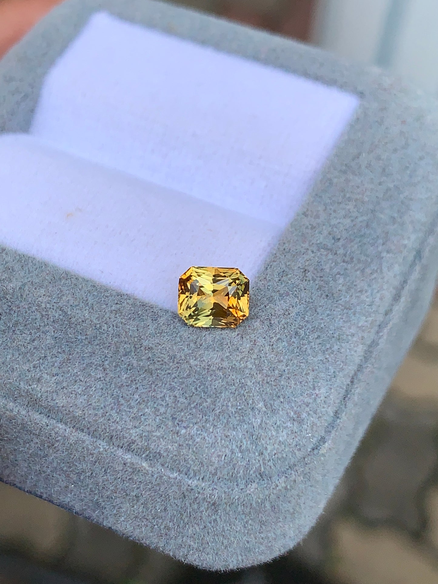 Le « Saphir bicolore • 1,10 ct » de Bonnot Paris est une magnifique pierre jaune rectangulaire aux angles arrondis, présentée avec élégance dans un écrin gris à l'intérieur en tissu blanc. Ses facettes rayonnent à la lumière naturelle, soulignant son exceptionnelle pureté.