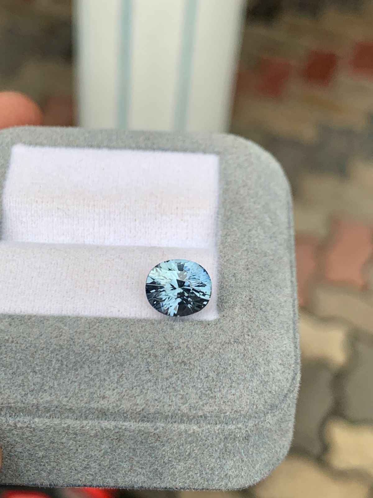 Un saphir gris bleu rond de 3,10 carats de Madagascar de Bonnot Paris scintille sur une base coussinée blanche à l'intérieur d'un coffret en velours gris, avec des pavés terreux flous en arrière-plan pour souligner son élégance.