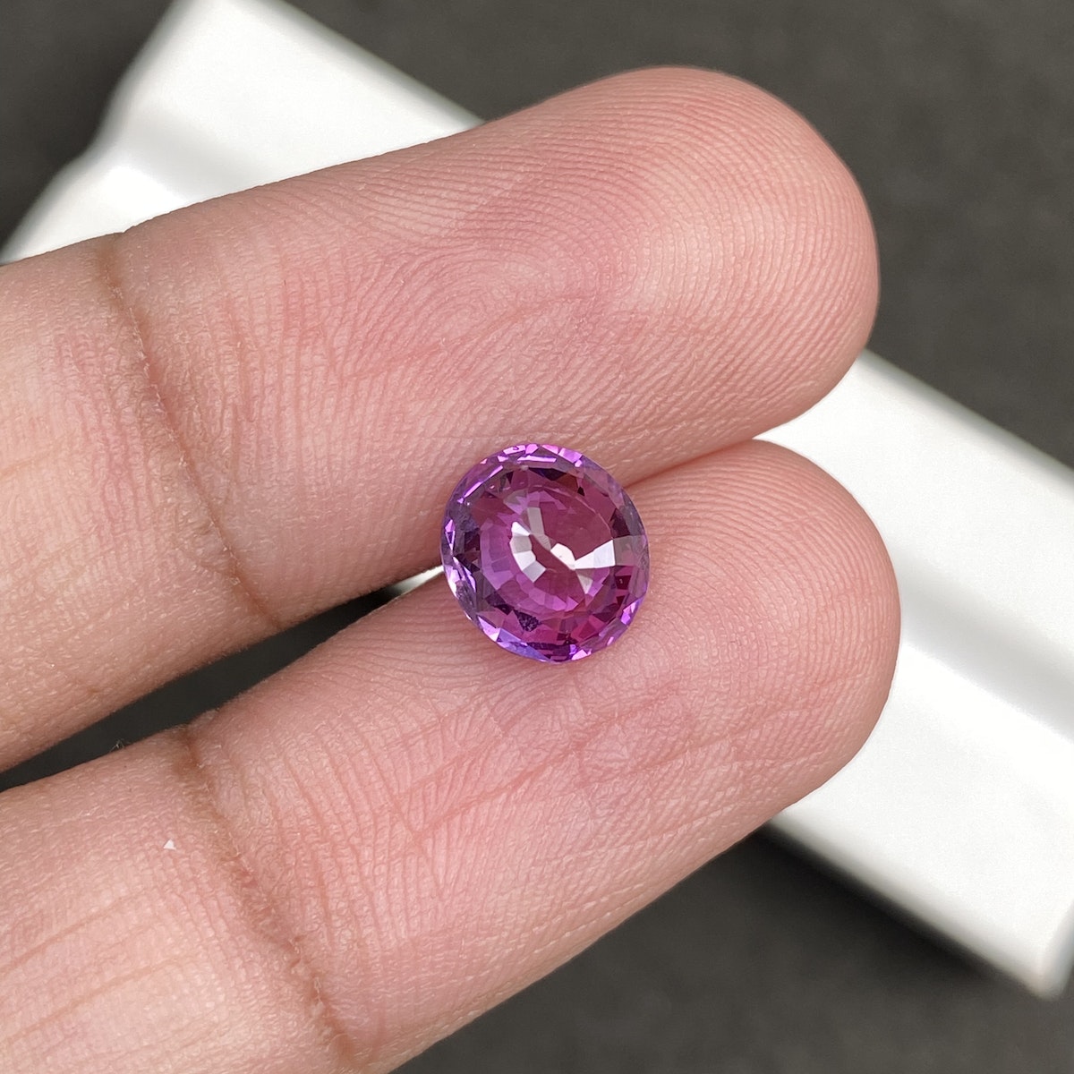 Une main tient le « Saphir Rose · 2,03 ct » de Bonnot Paris, une petite pierre violette ronde et facettée qui scintille sous la lumière. L'arrière-plan est flou et grisâtre.