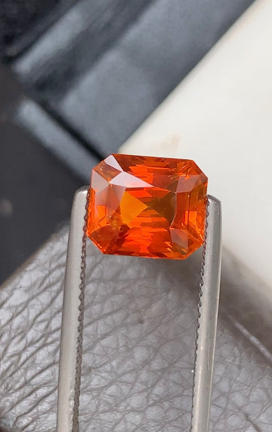 Tenu par une pince en argent, le Saphir Orange • 4,53 ct de Bonnot Paris éblouit par sa teinte vive et ardente et sa surface brillamment facettée reflétant la lumière sur un fond brun texturé et blanc lisse.