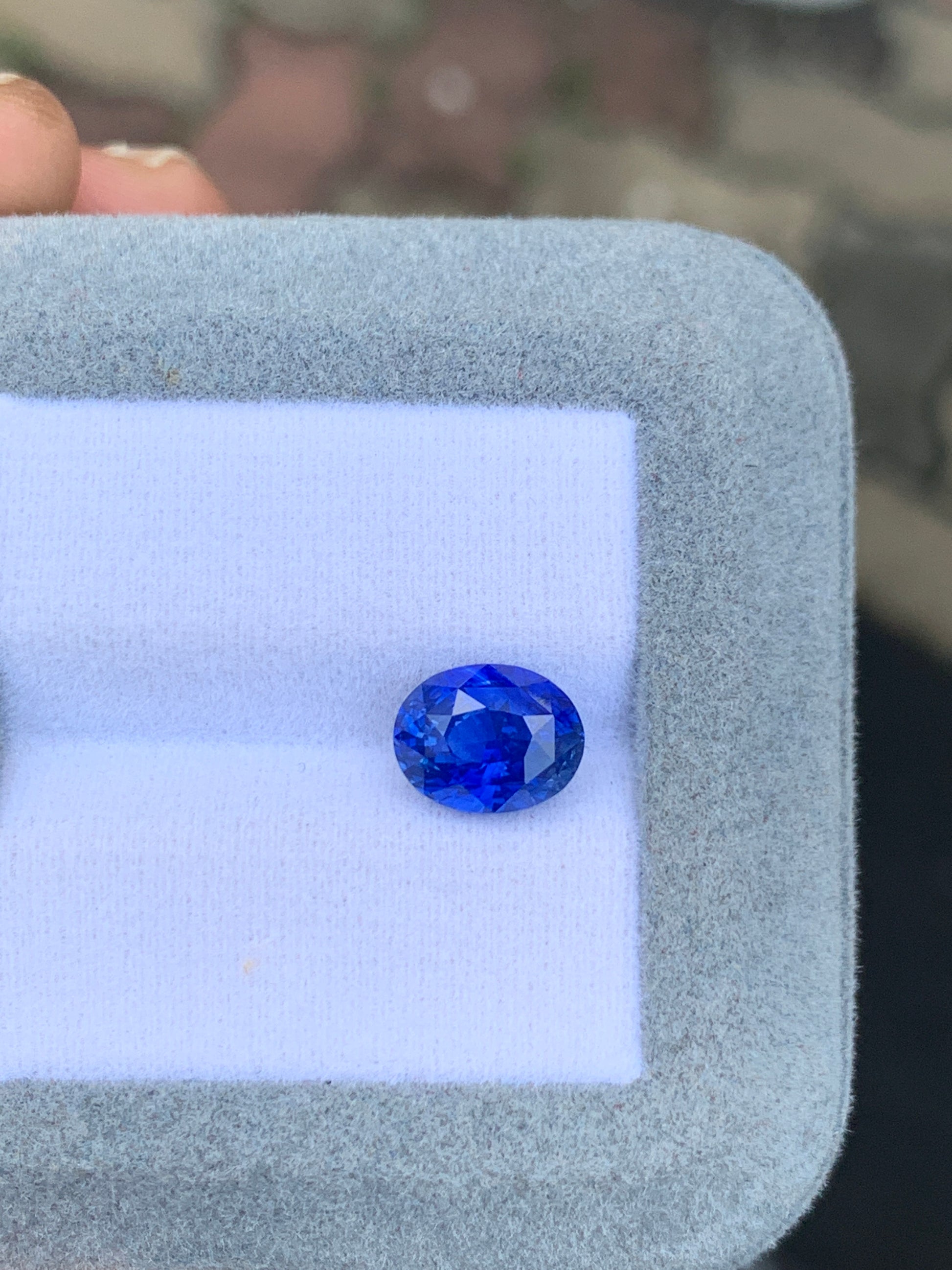 Le Saphir bleu de 3,55 carats de Bonnot Paris, ressemblant à une pierre précieuse du Sri Lanka, repose dans une vitrine texturée gris clair, mettant en valeur sa couleur bleu vif et sa clarté - une véritable gemme d'exception.