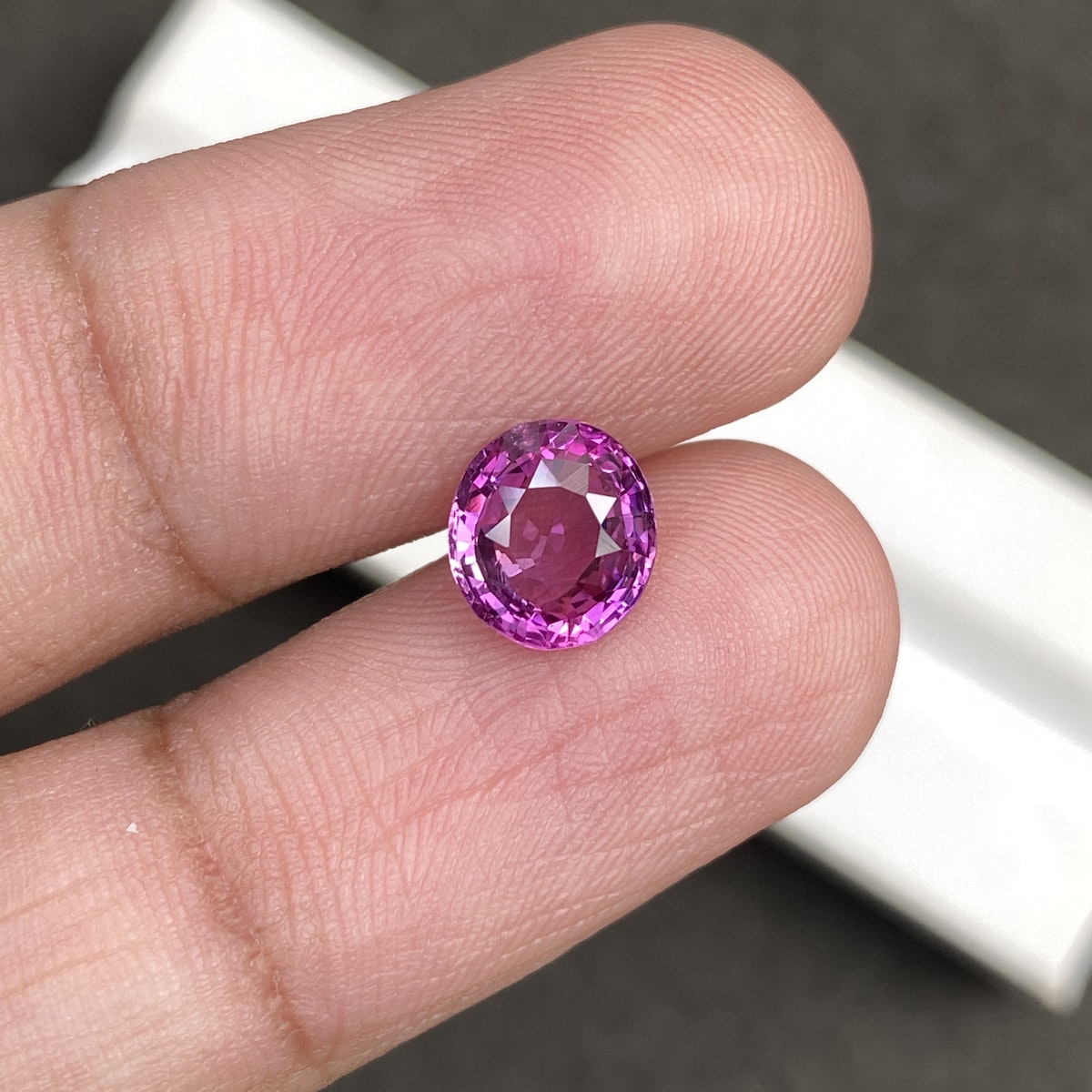 Une personne tient entre son pouce et son index le Saphir Rose · 2,03 carats, une pierre précieuse taille ronde étincelante de Bonnot Paris. L'arrière-plan flou met en valeur la couleur rose éclatante et les facettes de la pierre.