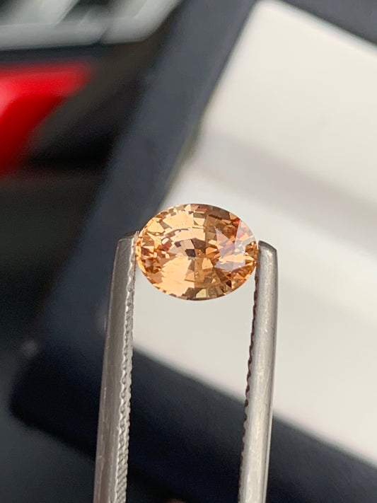 Délicatement tenue par une pince à épiler, la radieuse pierre précieuse orange taille ovale de 1,2 ct de Bonnot Paris Saphir Pêche en provenance de Madagascar scintille sur un fond flou, mettant en valeur sa clarté et ses facettes.