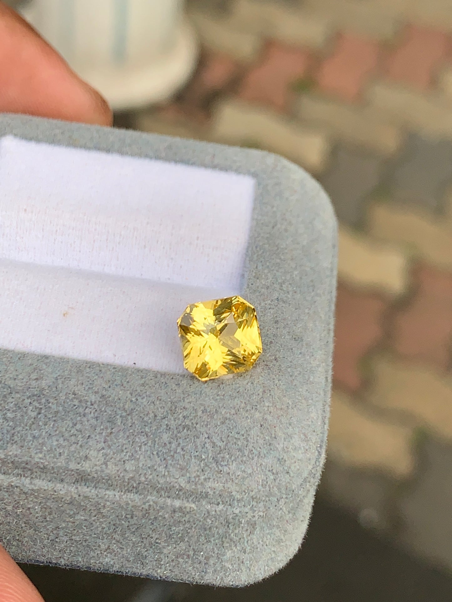 Une personne tient un coffret à bijoux gris ouvert contenant un Saphir jaune Bonnot Paris de 4,00 carats. L'arrière-plan floute artistiquement les pavés dans des tons gris et rouges, faisant écho au célèbre patrimoine gemmologique du Sri Lanka.
