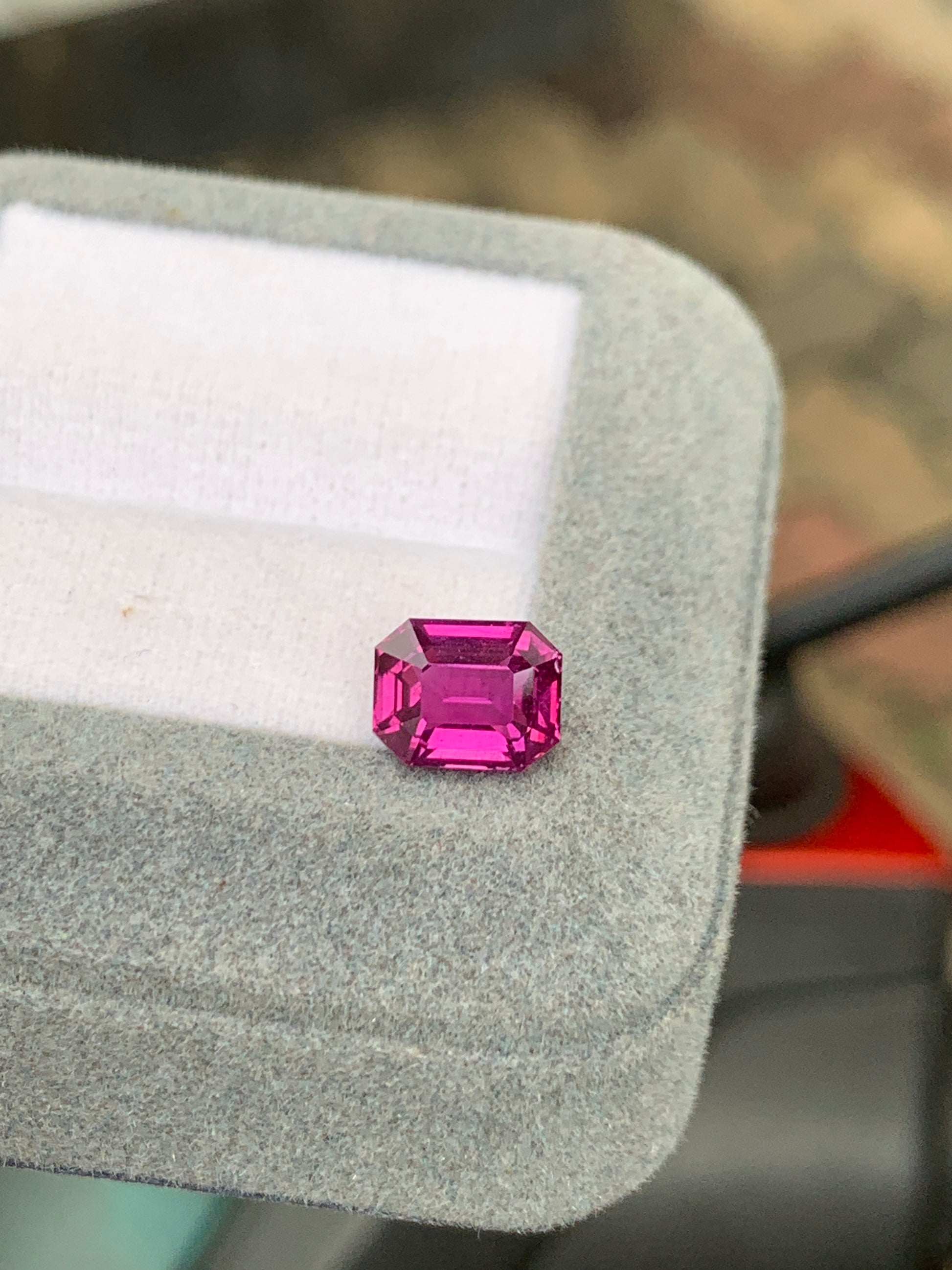 Un Saphir rose éclatant de 3,02 ct de Bonnot Paris, taille émeraude, repose sur un coussin gris doux dans un écrin ouvert. Le fond légèrement flou met en valeur la couleur riche et la surface facettée de la pierre précieuse.