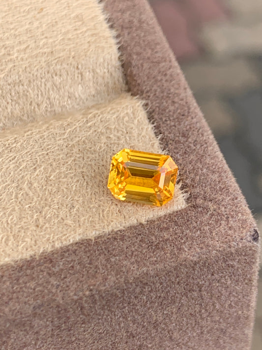 Un Saphir Jaune rectangulaire de 2,2 carats, taille émeraude, signé Bonnot Paris, repose sur une surface brun clair et floue. Ses facettes captent la lumière, créant un effet éblouissant rappelant la haute joaillerie sur un fond flou.