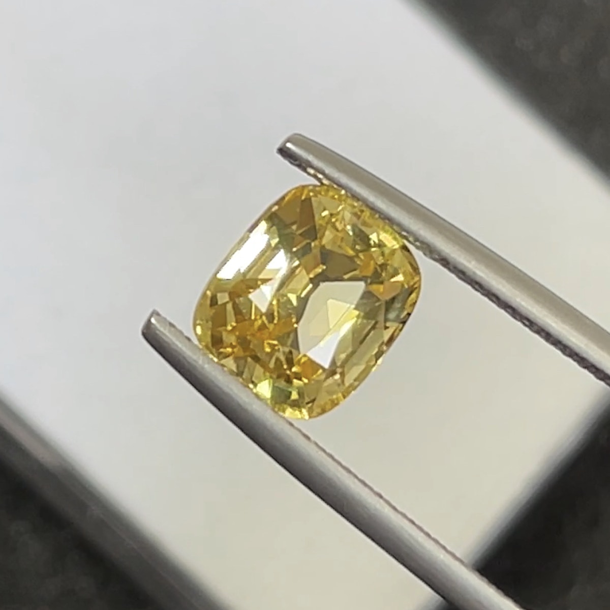Le Saphir Jaune · 3,82 ct de Bonnot Paris, une pierre précieuse jaune taille coussin facettée, est maintenu en gros plan par une pince en argent sur un fond neutre flou, soulignant sa beauté réfléchissante.