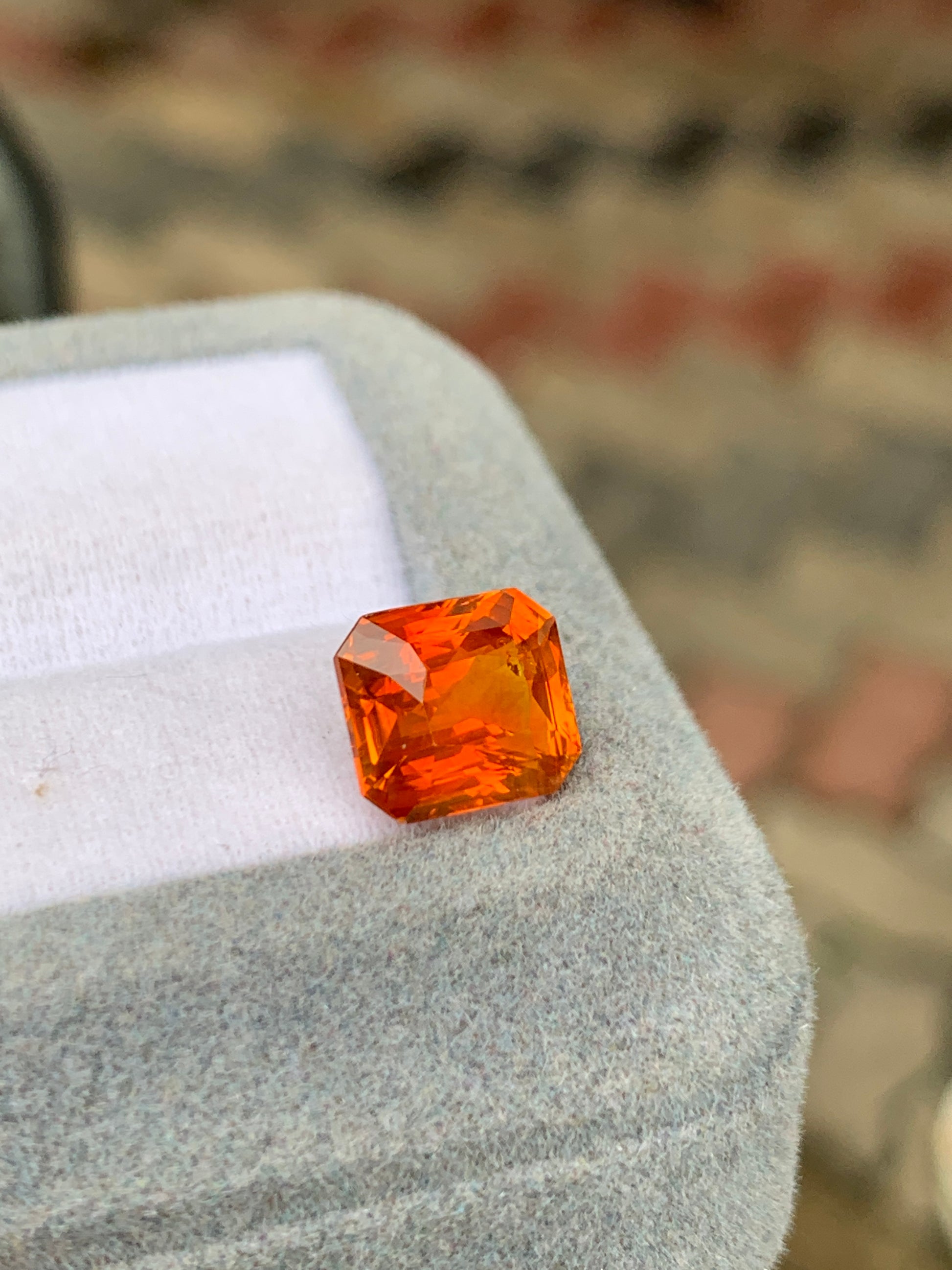 Un Saphir Orange taille émeraude vibrant (4,55 ct) de Bonnot Paris repose sur un coussin en tissu gris clair au sommet d'une boîte, avec un fond à motifs flous.