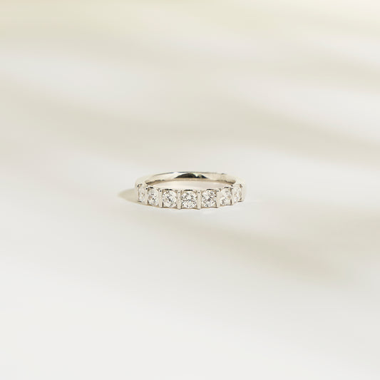 L'Alliance La Subtile de Bonnot Paris est une bague en argent sertie de pierres taille carrée, semblables à des diamants. Son éclat brillant évoque un luxe discret, issu d'une collection exquise. Elle repose sur une surface lisse et légère, avec des ombres douces et floues en arrière-plan.