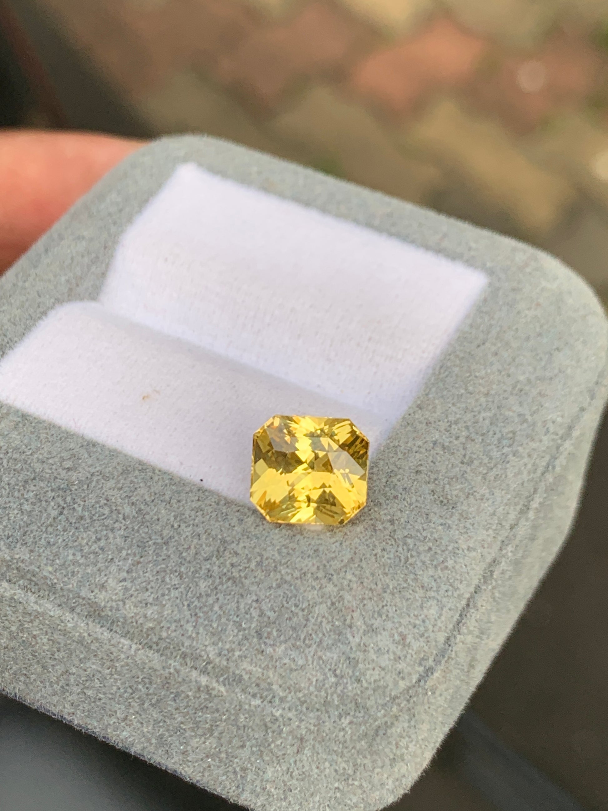 Une boîte à bijoux grise ouverte avec un intérieur blanc repose dans la main de quelqu'un, affichant un superbe saphir jaune taille octogonale de 4,00 ct de Bonnot Paris sur un fond flou, mettant en valeur la beauté naturelle des célèbres pierres précieuses du Sri Lanka.