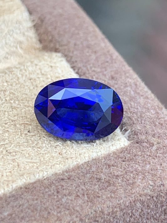 Gros plan sur le Saphir Bleu • 3,51ct taille ovale de Bonnot Paris qui scintille d'un bleu profond sur une surface texturée beige, sur fond brun flou.