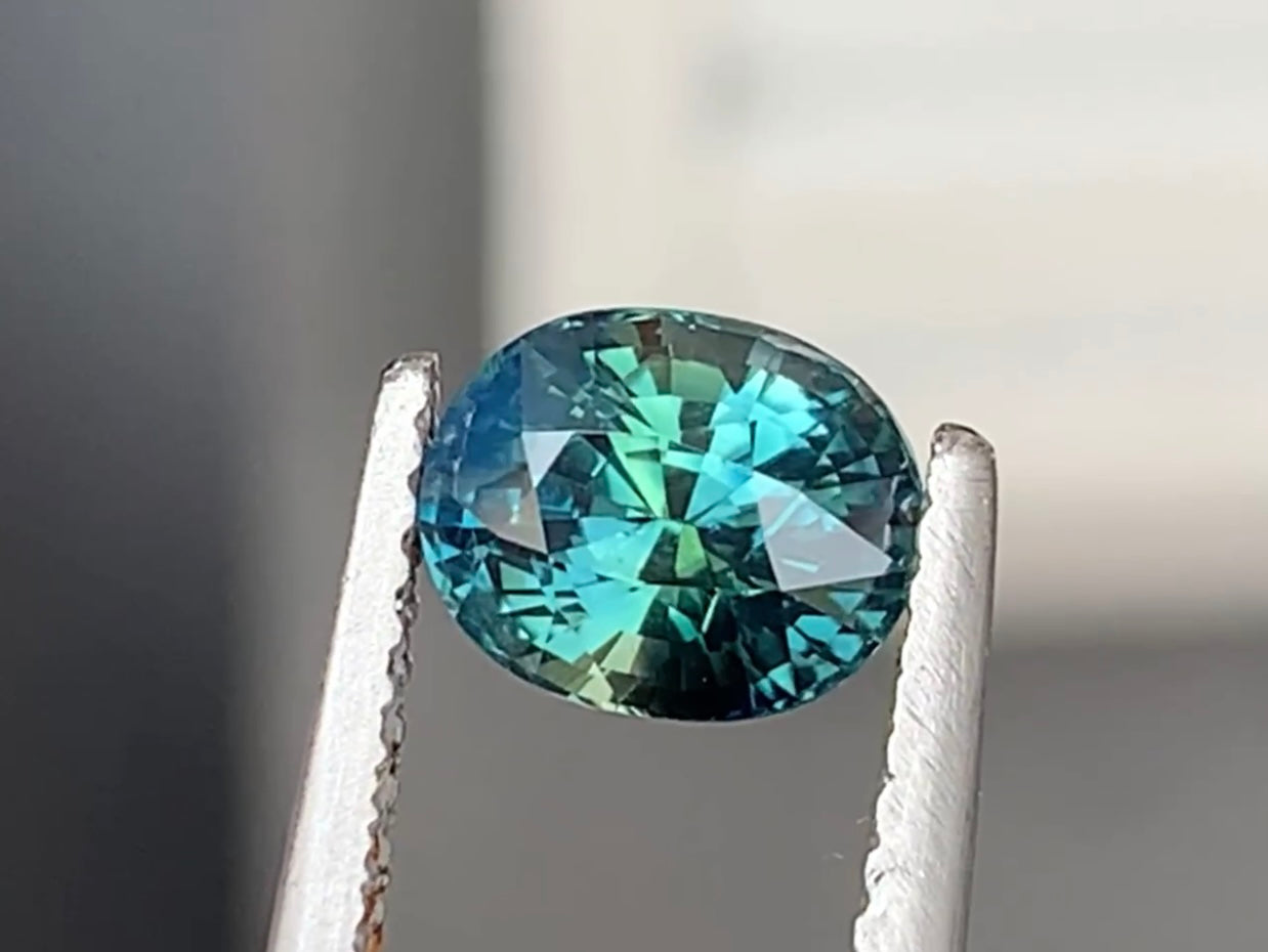Une pierre précieuse ronde et facettée de la collection « Saphir Teal • 1,6 ct » de Bonnot Paris est maintenue par une pince métallique. Sa surface scintille de nuances de bleu et de touches de vert sur un fond neutre et flou.