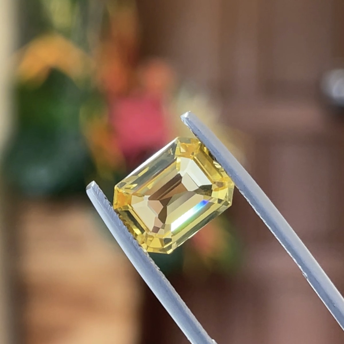 Une pierre précieuse jaune de 4,04 carats de la collection Saphir Jaune de Bonnot Paris est maintenue dans une pince métallique sur un fond flou de teintes vertes et brunes.