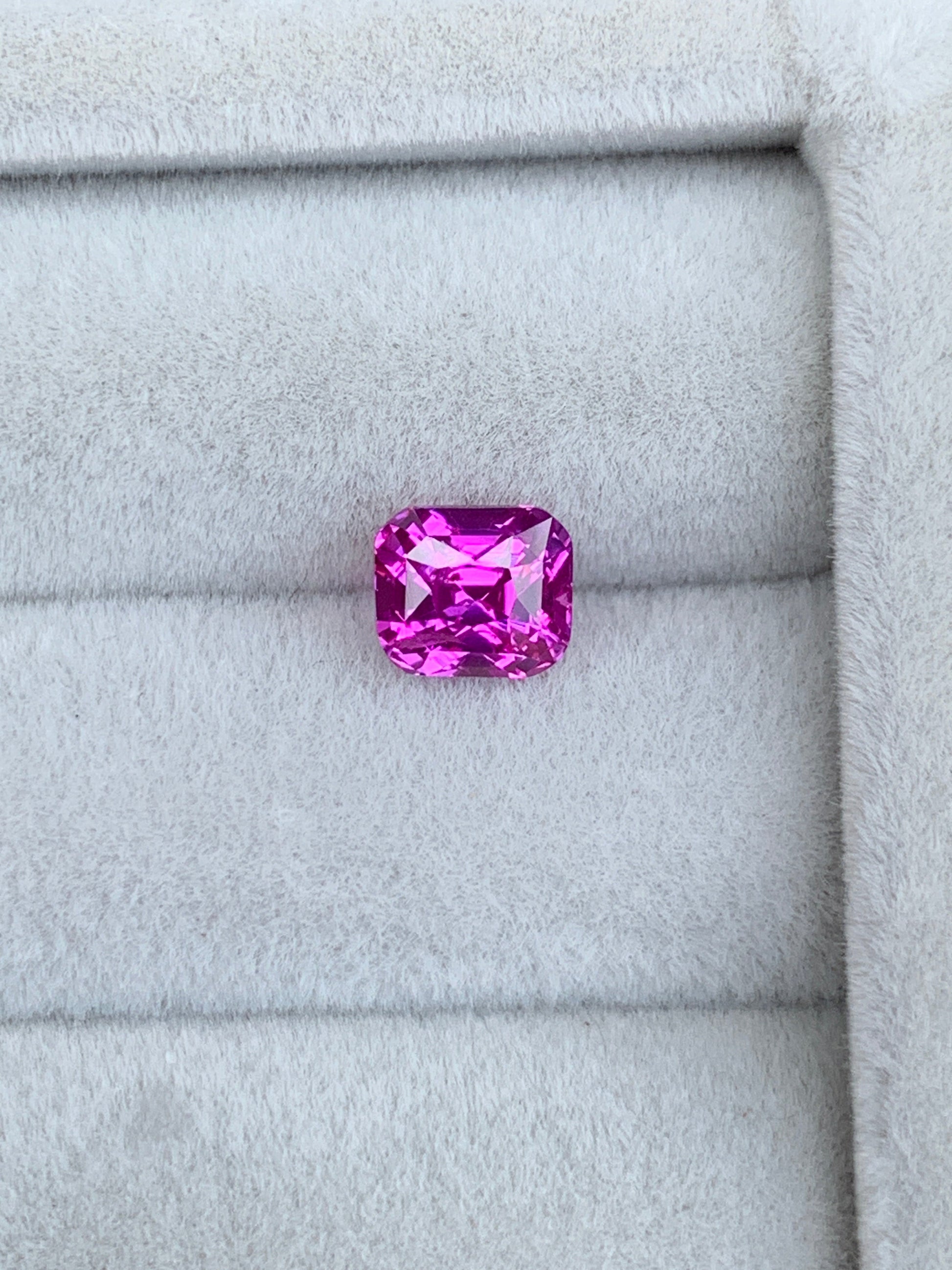 Un Saphir Rose éclatant de 1,6 carat signé Bonnot Paris repose sur un tissu gris doux, avec une taille rectangulaire aux bords biseautés. Ses facettes reflètent brillamment la lumière, sublimant sa couleur vive : un véritable bijou sur mesure.