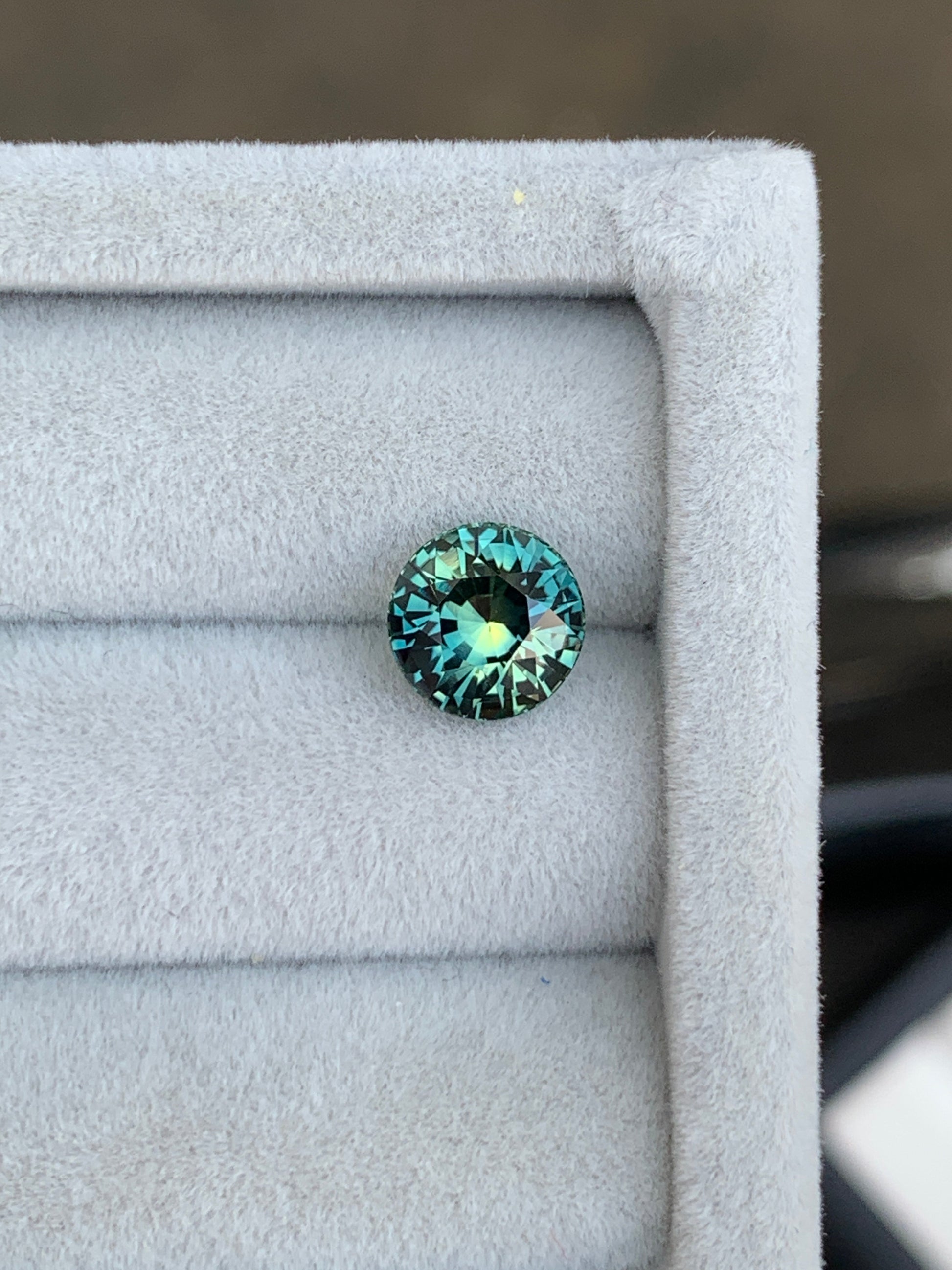 La pierre précieuse Bonnot Paris Saphir Teal • 2,55 carats de Madagascar scintille de reflets bleu sarcelle et aqua sur un fond gris doux, sa surface facettée évoquant l'élégance d'une exquise bague de fiançailles.