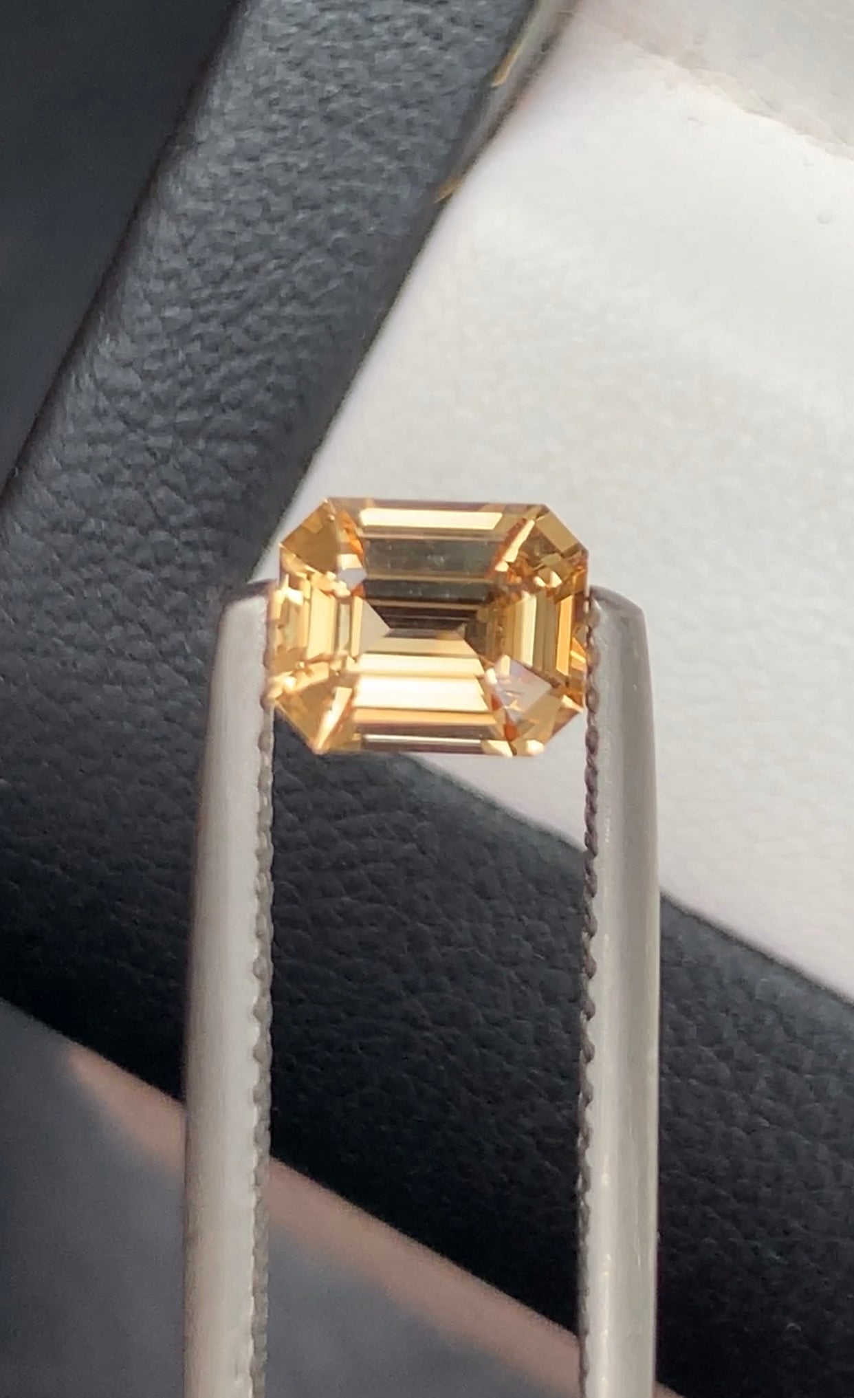 Un gros plan sur le « Saphir Pêche • 1,12ct » de Bonnot Paris révèle une pierre précieuse dorée taille émeraude maintenue par une pince métallique sur un fond sombre et texturé, évoquant un cadre professionnel.