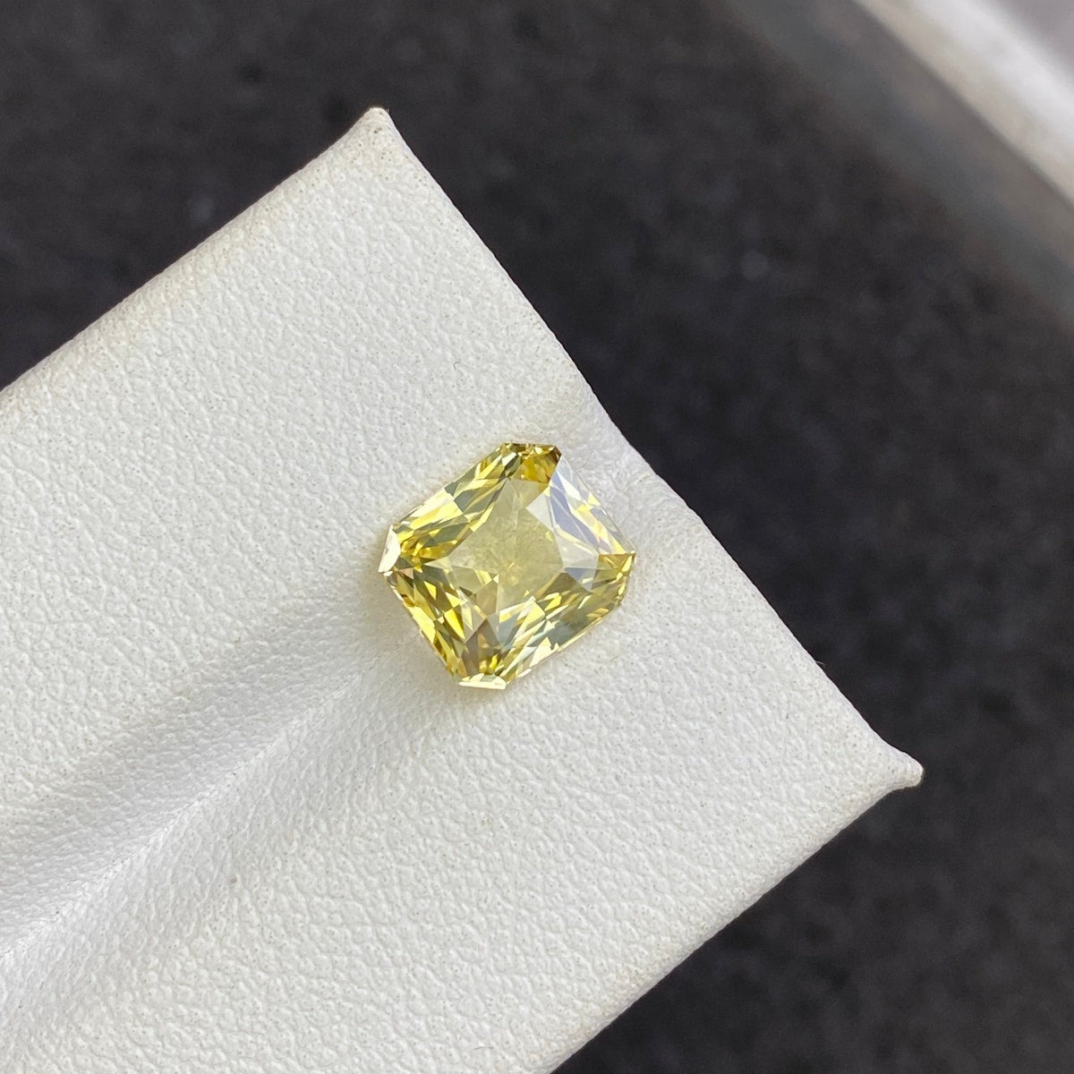 Le Saphir Jaune · 3,03 ct de Bonnot Paris, un diamant jaune taille radiant présenté sur un support blanc sur un fond sombre, met en valeur l'éclat et la clarté de la pierre précieuse.