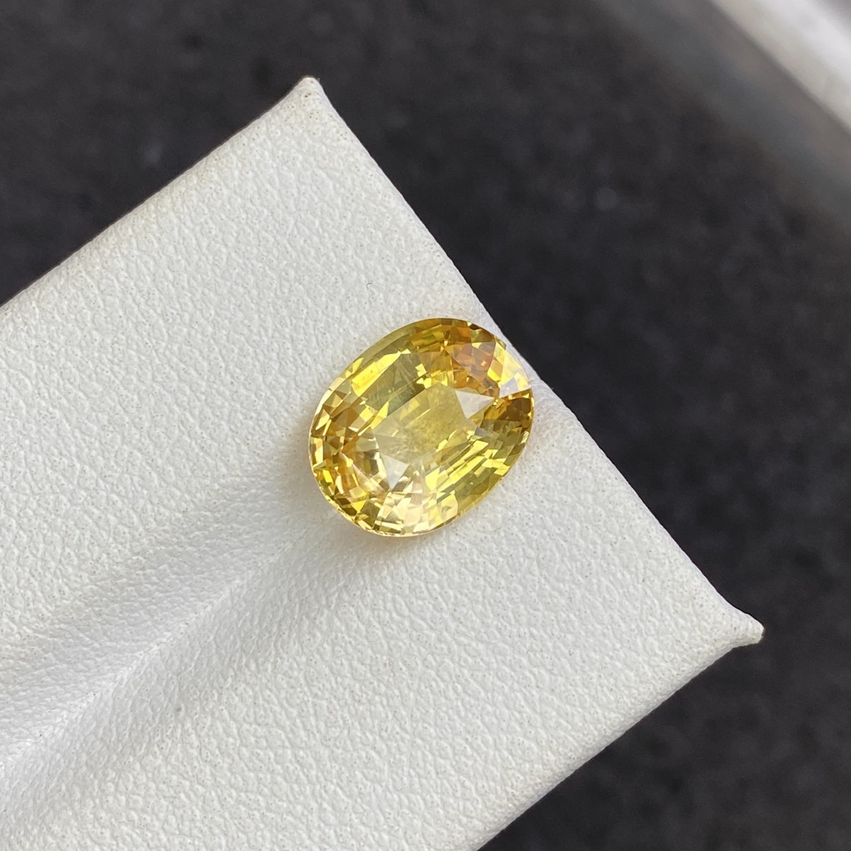 Une pierre précieuse jaune ovale étincelante de 4,5 carats, Saphir Jaune de Bonnot Paris, est présentée sur une surface blanche sur un fond sombre.