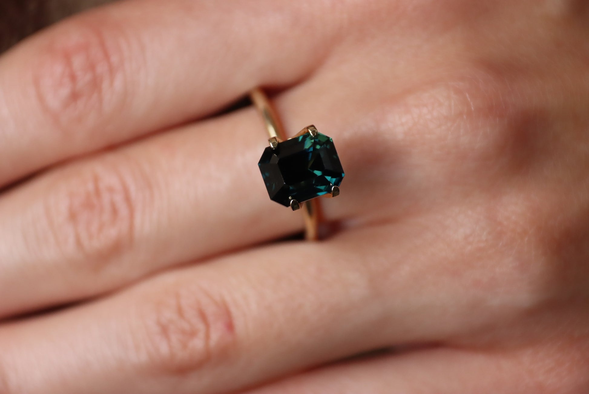 Une main porte le Saphir Teal · 4,04 carats de Bonnot Paris, orné d'une grande pierre rectangulaire vert foncé sertie sur or. Les doigts sont légèrement écartés, posés sur une surface.