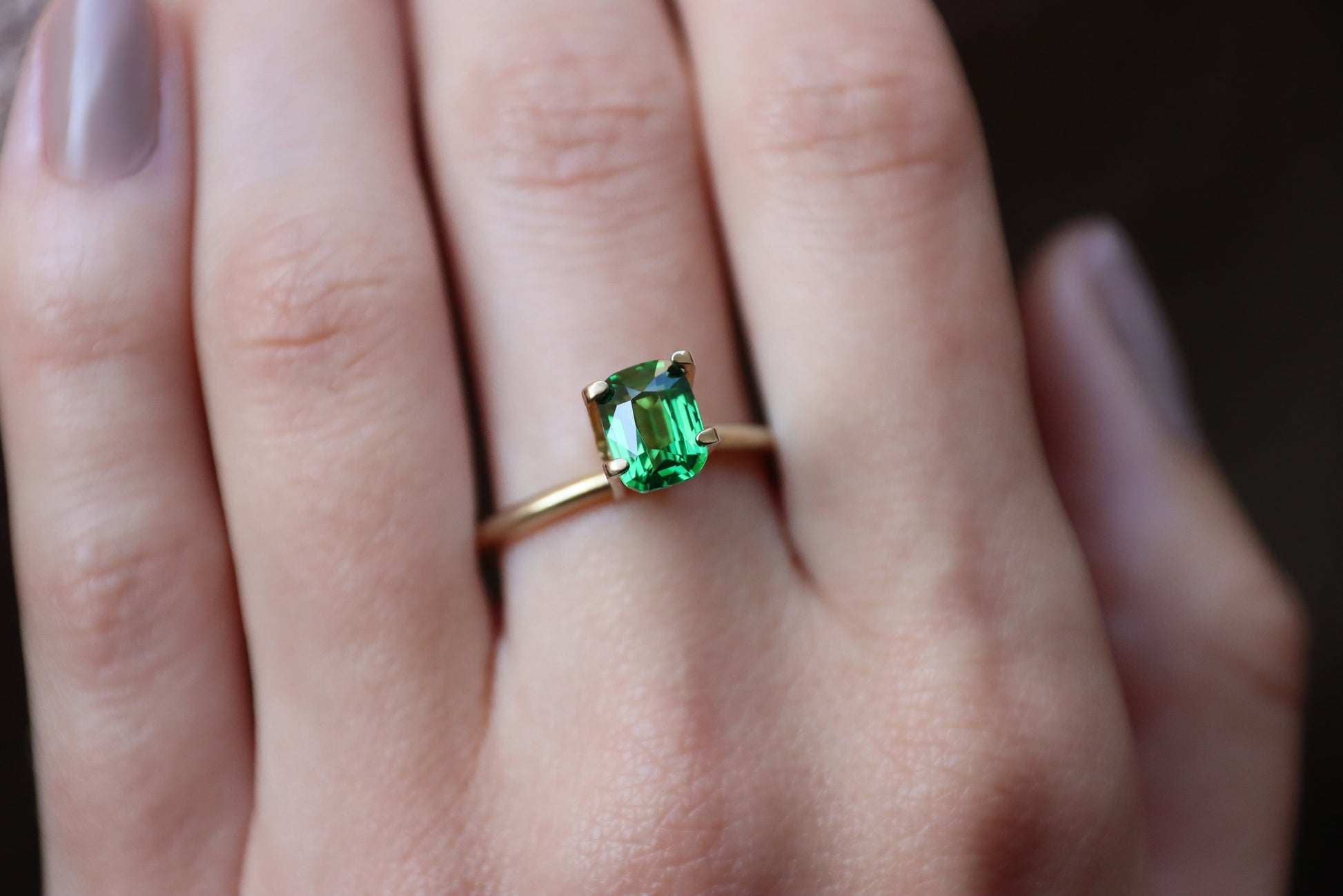 Gros plan d'une main portant la bague Bonnot Paris Grenat Tsavorite en or 1,54 carat ornée d'une pierre verte rectangulaire. Les doigts légèrement polis sont partiellement visibles sur un fond sombre et flou.