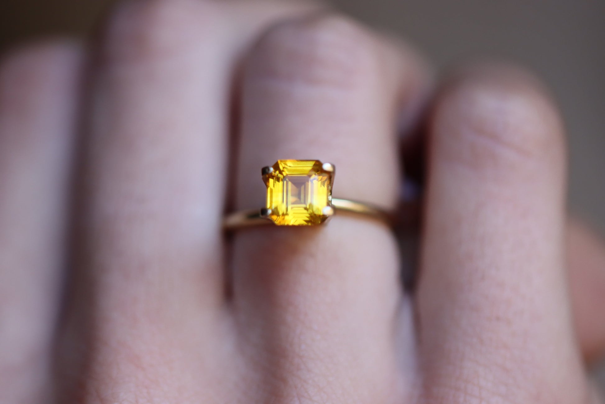 Une main met en valeur la bague Saphir Jaune · 2,04 ct de Bonnot Paris, comportant une pierre précieuse jaune taille carrée proéminente sertie d'or sur un fond flou.