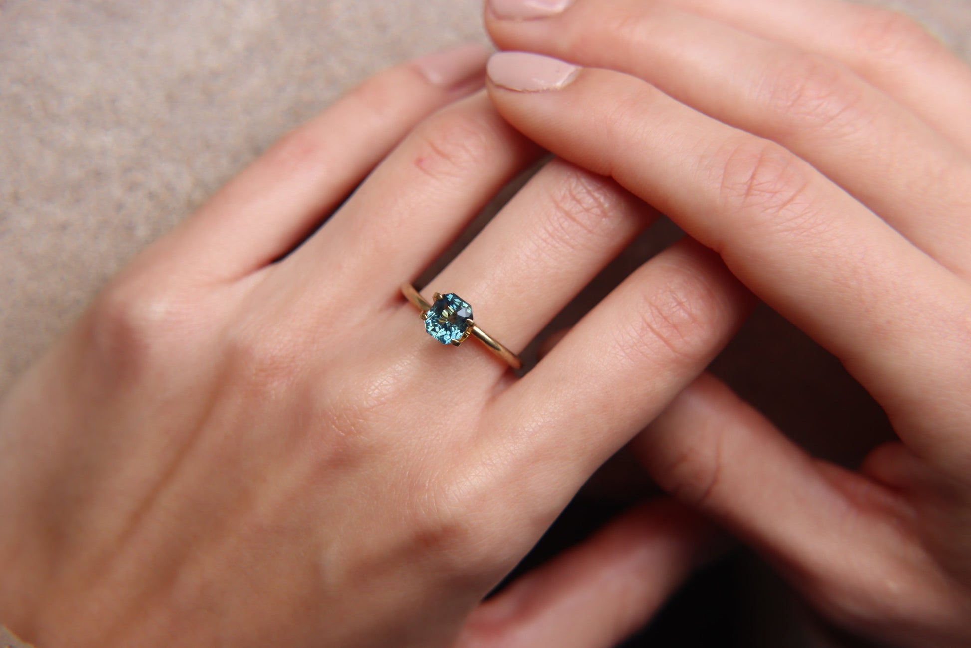 Gros plan d'une main ornée d'une bague Saphir Teal en or 1,6 carat de Bonnot Paris ornée d'une pierre ronde bleue. Les ongles de la personne sont élégamment manucurés avec un vernis neutre et clair sur un fond doux et flou mettant en valeur la bague.