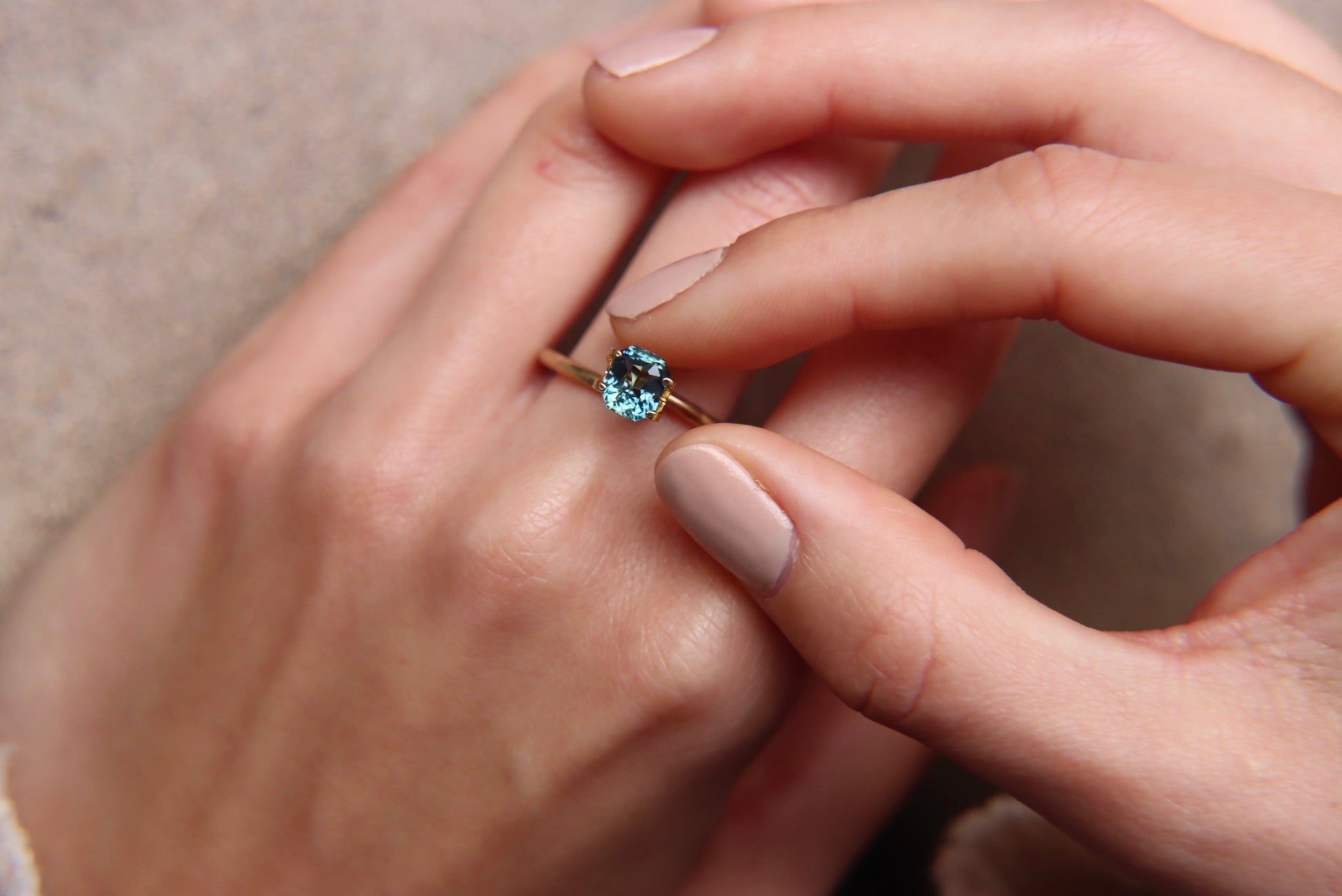 Un gros plan montre une main ornée d'une bague Saphir Teal en or 1,6 carat de Bonnot Paris, ornée d'une pierre bleue à l'annulaire. Une autre main effleure délicatement la bague, les ongles peints d'une teinte neutre, sur un fond doux et texturé.