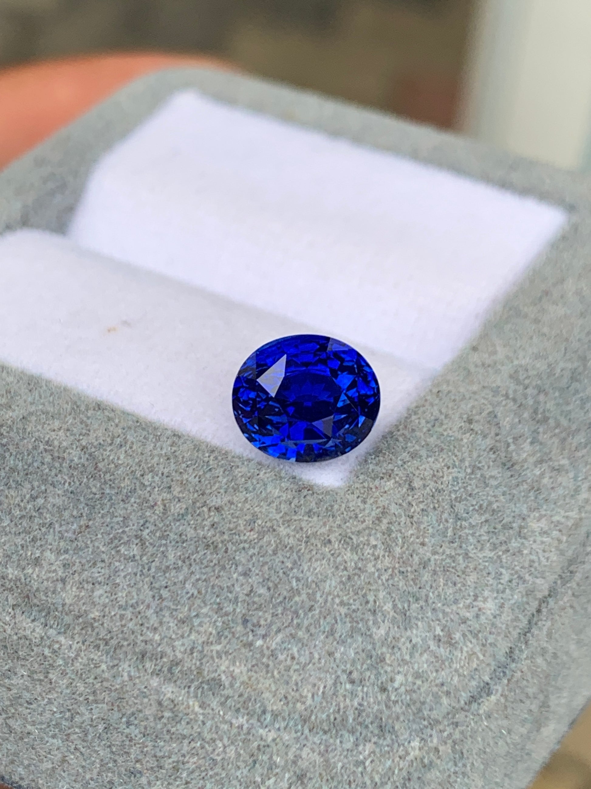 Le Saphir Bleu • 3,25ct de Bonnot Paris est une pierre précieuse ovale, d'un bleu vibrant, aux multiples facettes, présentée dans un écrin à bijoux blanc matelassé sur un extérieur en tissu gris.