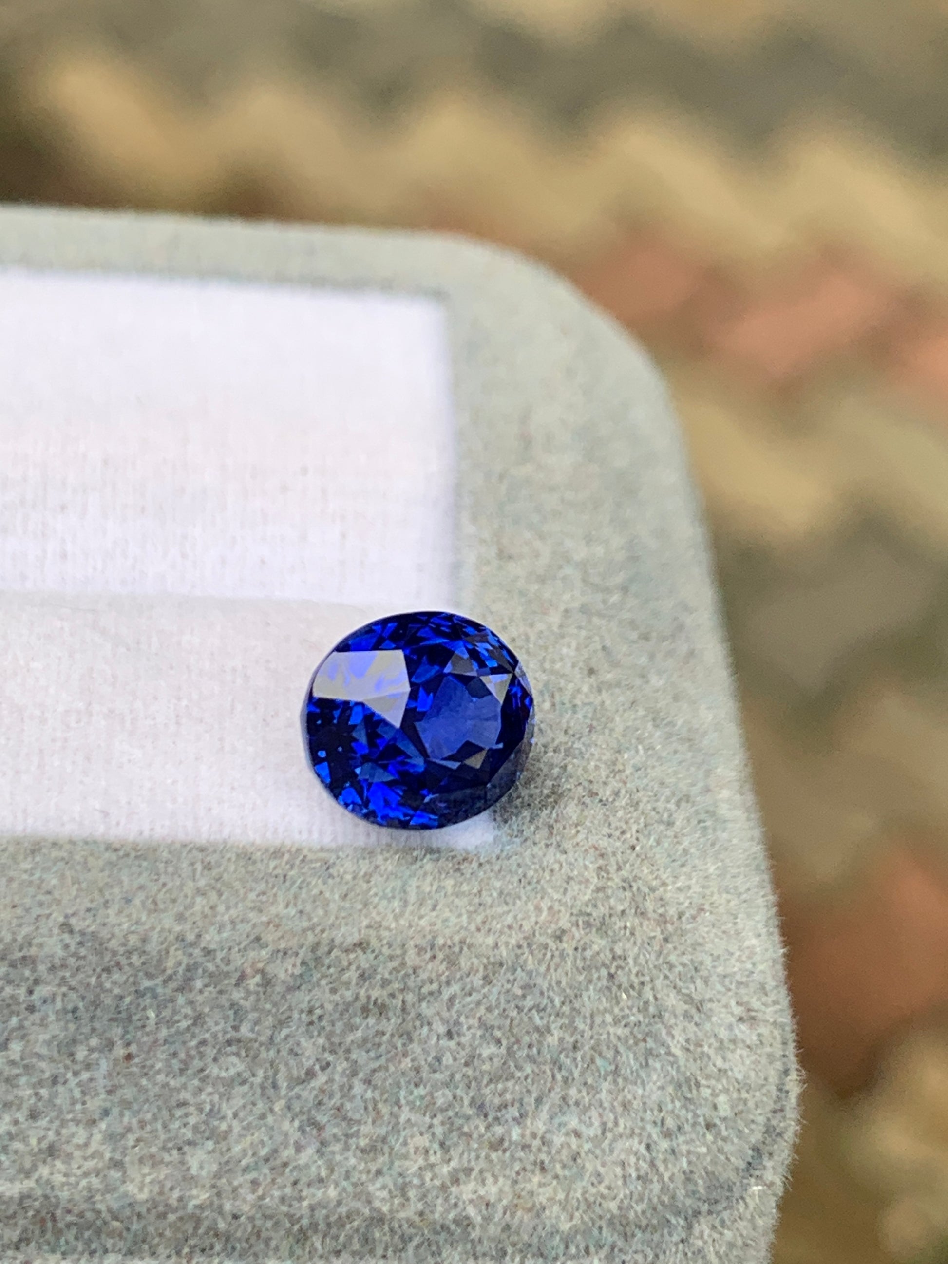 Le Saphir Bleu • 3,25ct de Bonnot Paris, une pierre précieuse bleue taille brillant, repose sur un tissu gris clair avec un fond à motifs en zigzag aux tons terreux.