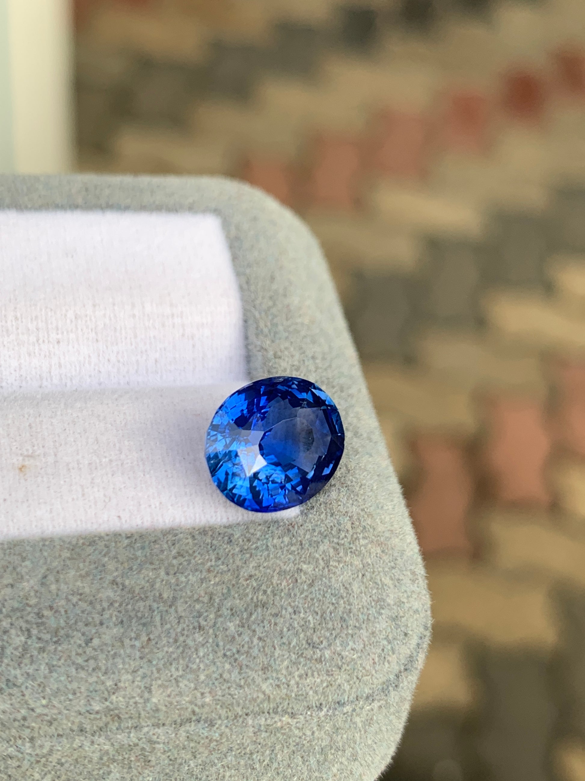 Gros plan d'un Saphir Bleu • 5 carats de [NOUVEAU] Bonnot Paris posé sur un tissu gris clair doux. La teinte bleu intense de la pierre précieuse ressort sur le fond légèrement flou aux motifs abstraits.