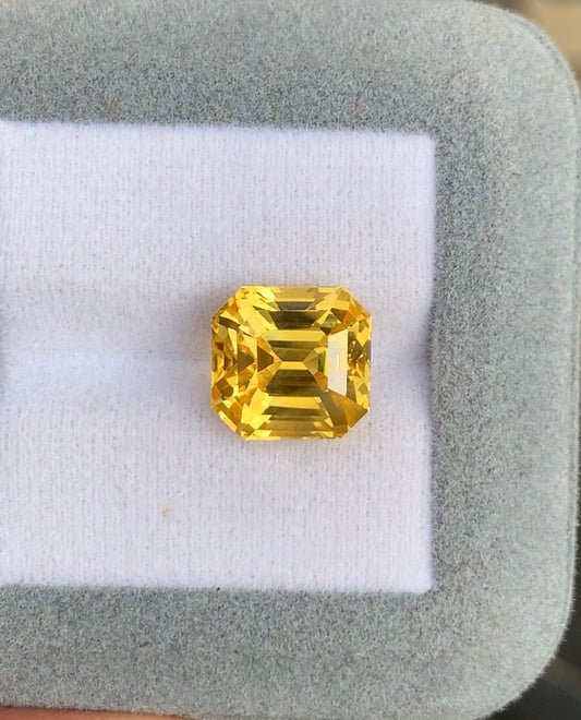 Une pierre précieuse jaune taille radiant est sertie sur un coussin blanc dans un écrin gris. Le Saphir Jaune de 10,15 carats de Bonnot Paris arbore une teinte vibrante, positionnée au centre pour mettre en valeur ses facettes et sa forme géométrique.
