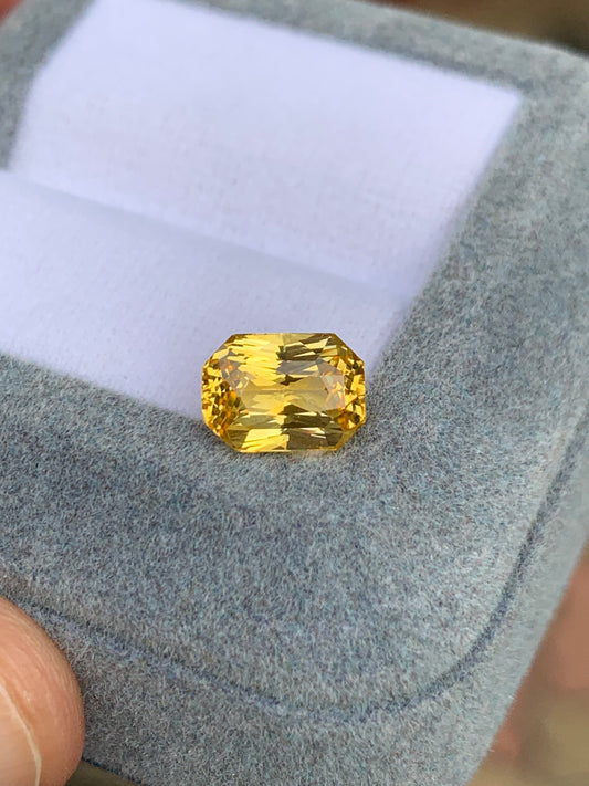 Un Saphir Jaune taille radiant de 2,53 ct de Bonnot Paris est présenté dans un écrin en velours gris à l'intérieur blanc, délicatement tenu entre les doigts.