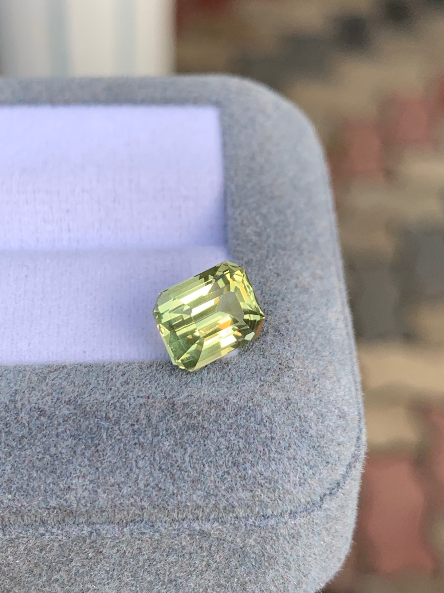 Le Saphir Jaune de 3,55 ct de [NOUVEAU] Bonnot Paris, une pierre précieuse taille radiant de Madagascar, est présenté sur une surface grise douce sous un angle pour mettre en valeur ses facettes et sa clarté sur un fond flou aux tons terreux.