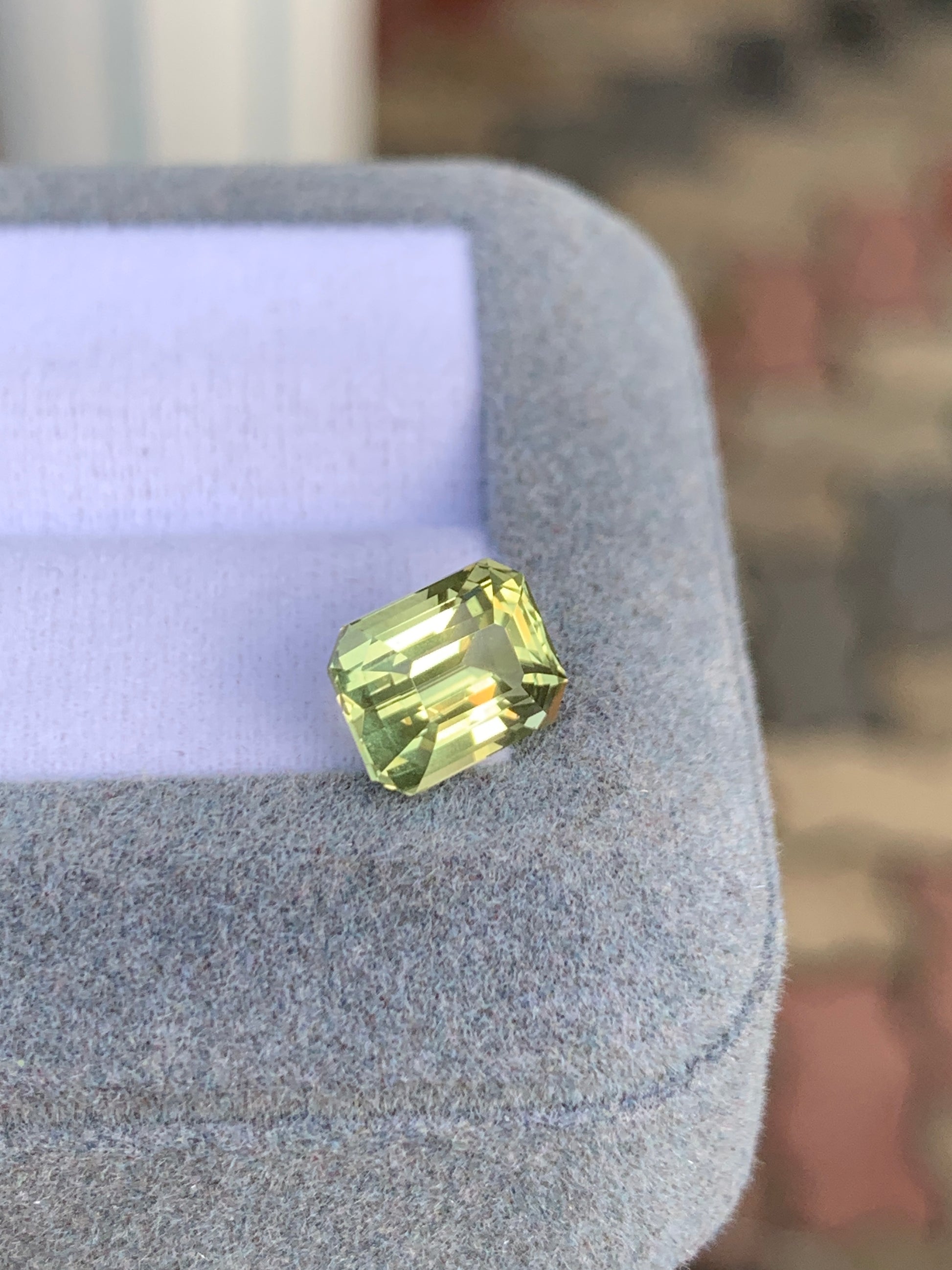Le Saphir Jaune de 3,55 ct de [NOUVEAU] Bonnot Paris, une pierre précieuse taille radiant de Madagascar, est présenté sur une surface grise douce sous un angle pour mettre en valeur ses facettes et sa clarté sur un fond flou aux tons terreux.