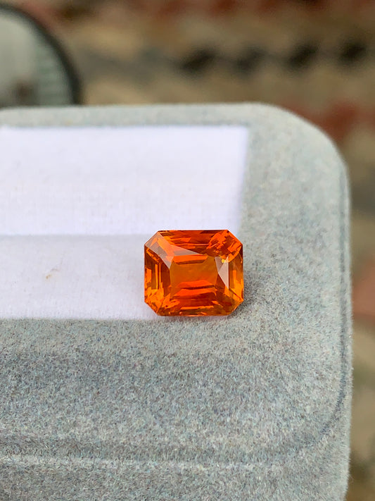 Le Saphir Orange de 4,55 carats de Bonnot Paris est présenté sur une surface grise matelassée. Sa teinte vive et flamboyante et ses facettes polies reflètent magnifiquement la lumière, faisant ressortir ce joyau précieux sur un fond légèrement flou.