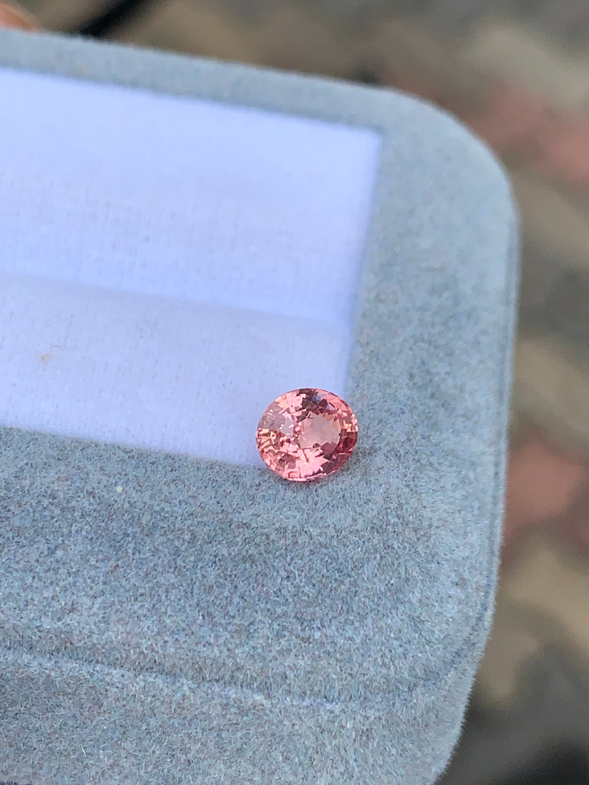 Le Saphir Padparadcha • 1,07 ct de [NOUVEAU] Bonnot Paris, une pierre précieuse rose ronde et scintillante, repose sur une boîte à bijoux gris clair avec un intérieur blanc sur un fond flou.