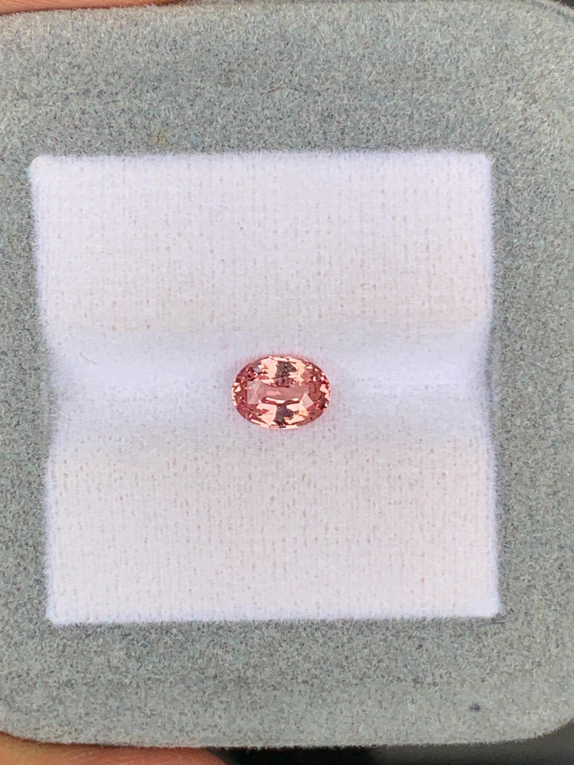Une petite pierre précieuse rose ovale de 1,02 ct de Madagascar, rappelant le rare Saphir Padparadscha de [NOUVEAU] Bonnot Paris, est présentée sur une surface coussinée blanche à l'intérieur d'un boîtier gris.
