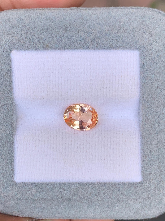 Un Saphir Padparadscha rond et facetté (2,04 carats) de Bonnot Paris est présenté sur un tissu blanc dans un support bleu-gris clair. Le fond flou met en valeur cette pierre précieuse exquise.