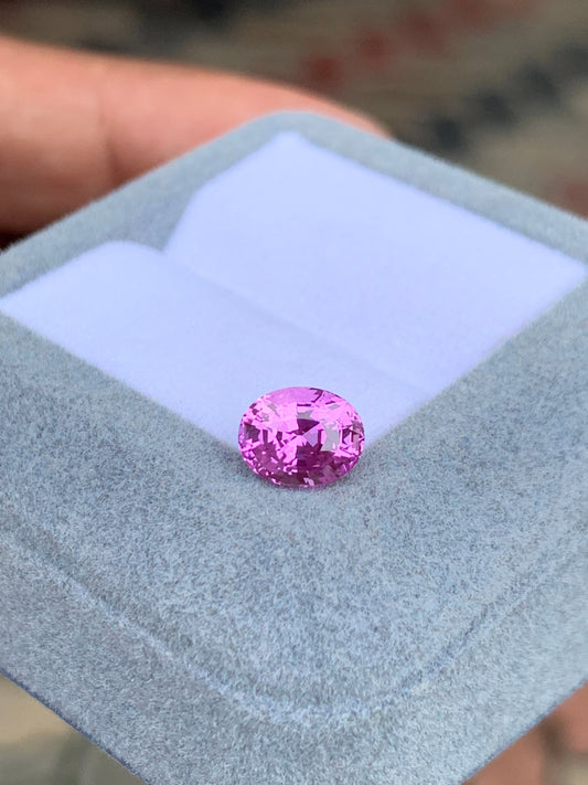 Une pierre précieuse Saphir Rose éclatante de Bonnot Paris, pesant 2,04 carats, est présentée sur un couvercle gris clair d'écrin, tenu par une main. Les facettes complexes de cette pierre ovale reflètent brillamment la lumière sur le tissu doux du fond.