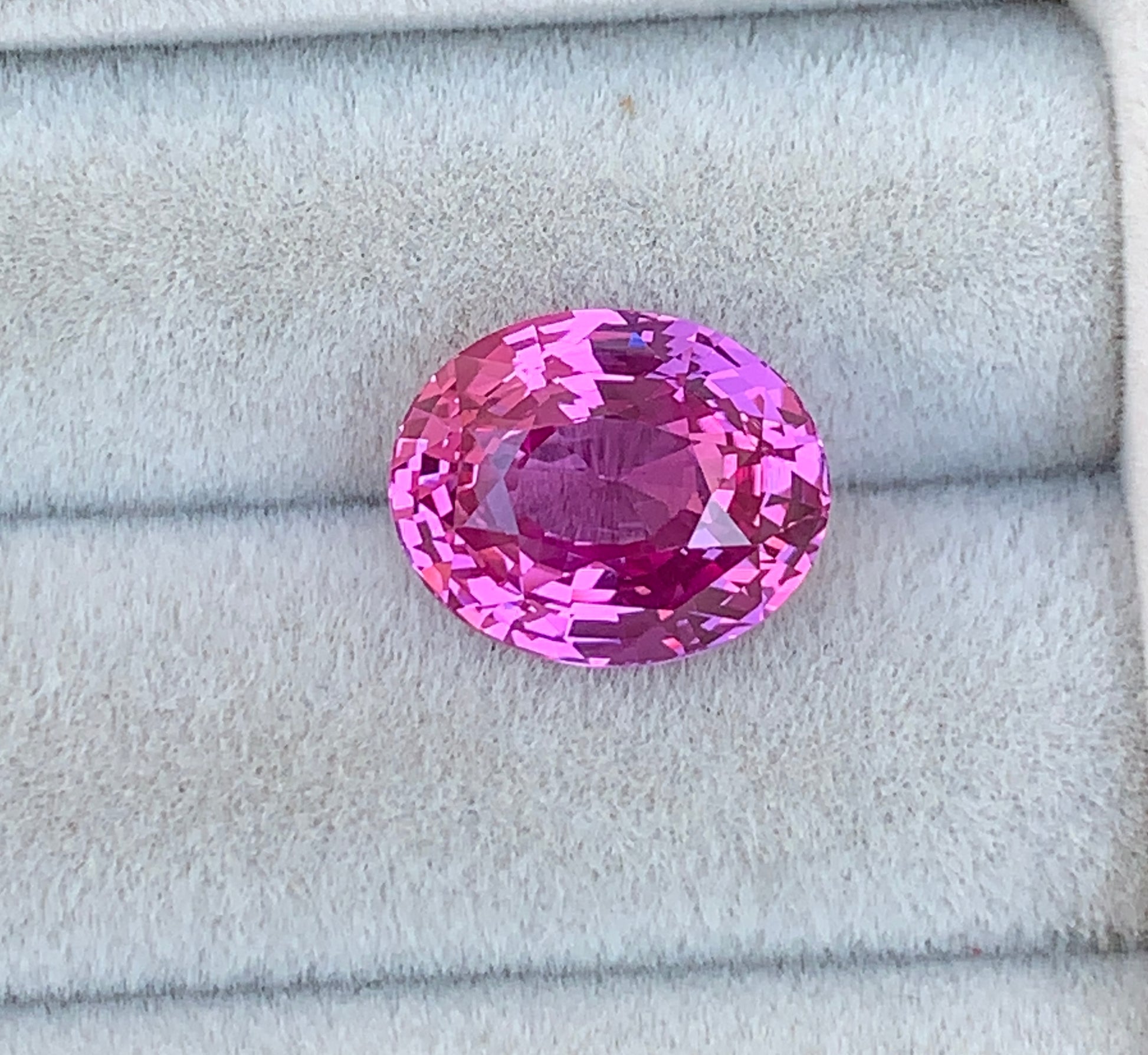 Un saphir rose ovale scintillant de 7,05 ct repose sur un fond de velours gris moelleux, mettant en valeur sa couleur vibrante et ses facettes complexes, un véritable témoignage de la rareté exceptionnelle de [NOUVEAU] Bonnot Paris.