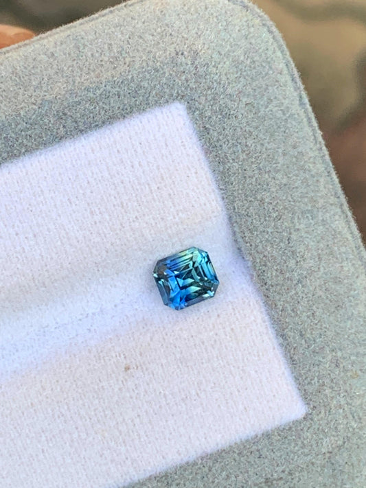 Le Saphir Teal • 1,10 ct de Bonnot Paris, avec sa taille coussin carrée, évoque la magnifique pierre précieuse de Madagascar. Présenté sur un tissu clair dans un écrin gris, il met en valeur des variations de couleurs éclatantes et des facettes scintillantes.