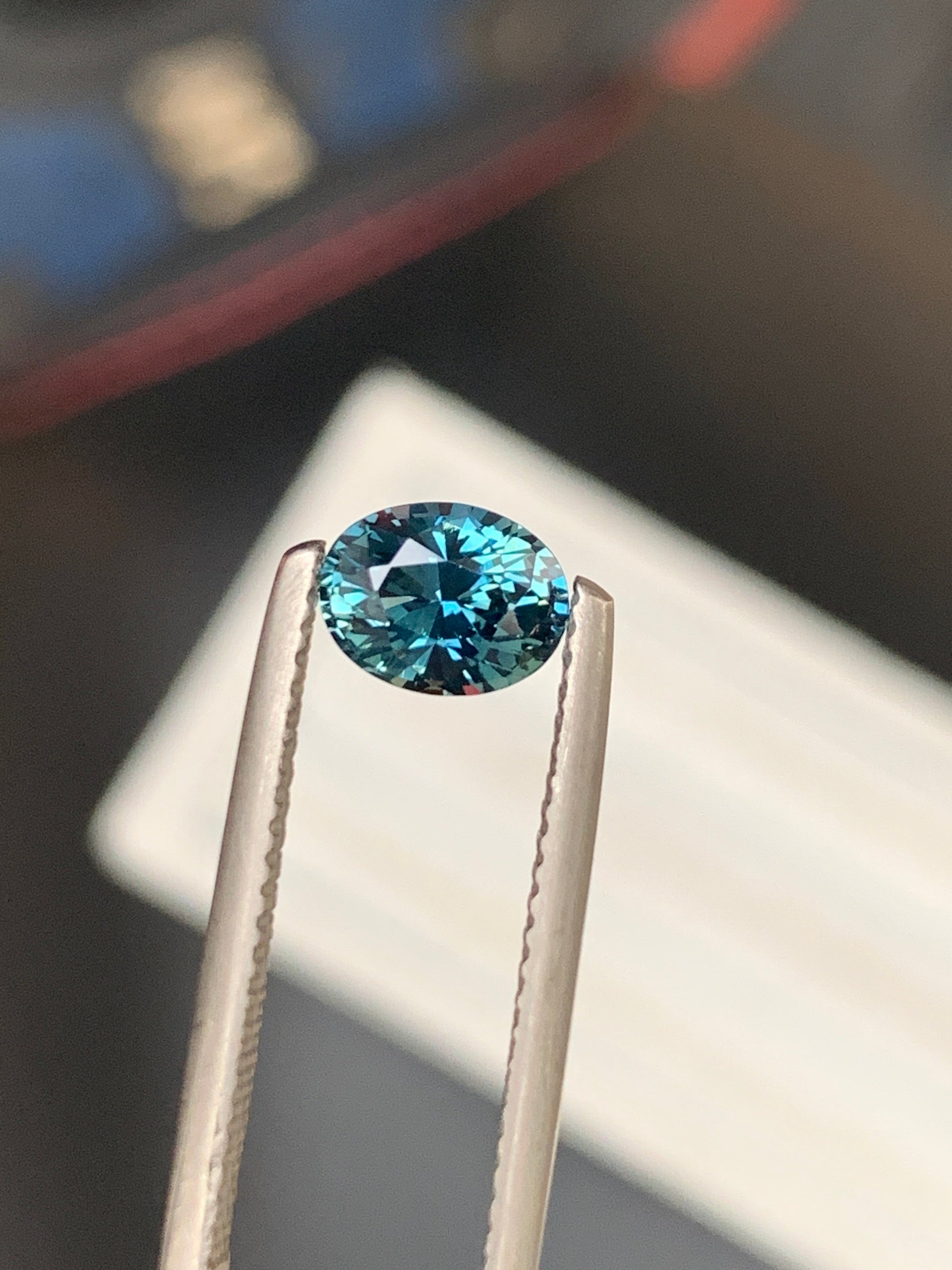 Une pince à épiler tient la pierre précieuse Saphir Teal • 1,25 carat de Bonnot Paris sur un fond flou. Sa teinte turquoise profonde et vibrante et ses facettes réfléchissantes capturent magnifiquement la lumière.