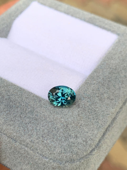 Le Saphir Teal (1,25 ct) de Bonnot Paris scintille de facettes complexes alors qu'il repose dans une boîte à bijoux en tissu gris avec un intérieur blanc, maintenue ouverte pour mettre en valeur la pierre précieuse.
